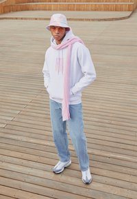 Grå hettegenser med rosa grafikk, lyseblå jeans, rosa skjerf drapert over skuldrene, hvite og grå joggesko, og en pastell-farget bøttehatt.