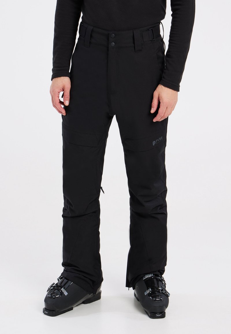 Homme portant un pantalon de neige noir et des chaussures de ski noires, debout sur un fond blanc.