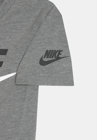 T-shirt cinza de manga curta feito de algodão. Apresenta um grande logotipo preto da Nike na manga e uma faixa branca atravessando o peito.