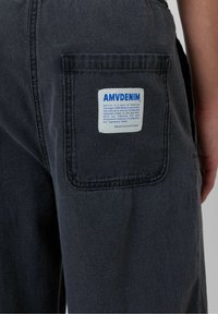 Pogled od blizu na zadnji žep temno modrih kavbojk z belo pravokotno oznako blagovne znamke AMVDENIM, ki je prišita na žepu.