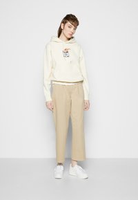 Cremefarbener Hoodie mit Kängurutasche und grafischem Detail, kombiniert mit beigen weiten Hosen und weißen Sneakern. Geschmeidige, lässige Stoffstrukturen.