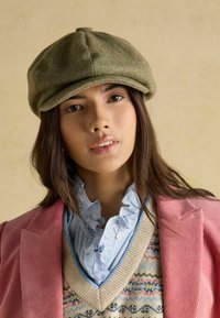 Zelená šiltovka so vzorom rybej kosti a zaoblenou korunou, nosená s ružovým šnurovacím sakom, vrstvenou modrou košeľou s volánmi a vzorovanou pletenou vestou.