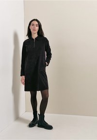 Czarna sukienka z zamkiem, uszyta z miękkiego materiału, z długimi rękawami i kołnierzykiem. Połączona z czarnymi rajstopami i butami wojskowymi, na neutralnym tle.