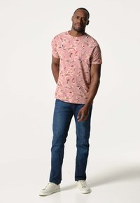 Korte mouwen roze t-shirt met een bladerenpatroon, gecombineerd met blauwe jeans en witte sneakers. Het shirt heeft een ronde halslijn en een klein logo op de arm.