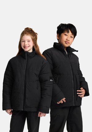 Smiling girl and boy wearing black puffer jackets and black pants standing side by side against a plain white background.