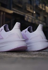 Двойка бели и светло лилави бягащи обувки Adidas Ultraboost на открит настил с размит градски фон.
