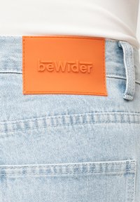 Πορτοκαλί δερμάτινο ταμπελάκι με την ένδειξη «bewider» ανάγλυφη στη ζώνη τζιν τζιν με ανοιχτό γαλάζιο χρώμα που φοριούνται με λευκό τοπ.