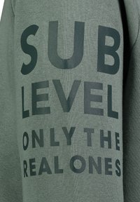 Grüne Sweatshirt mit fettem schwarzen Text, der "SUB LEVEL ONLY THE REAL ONES" lautet. Hergestellt aus weichem Stoff mit strukturierter Oberfläche.