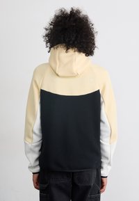 Hooded sweatshirt melnā, gaiši dzeltenā un baltā krāsā. Iezīmē krāsu bloku dizainu, mīkstu materiālu un elastīgas piedurknes.