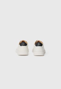 Hvide sneakers med en glat tekstur, der har en sort hælaccent og en tan inderside. Gummisåler, minimalistisk design og syninger.