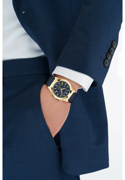 Reloj dorado con un dial analógico azul y correa de goma, llevado en una mano con un traje de color azul marino, que presenta un cuello con muescas y puño abotonado.