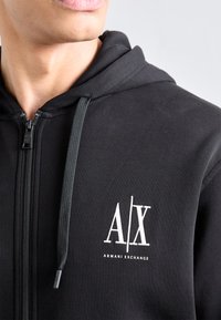 Juodas užtrauktukas megztinis, pagamintas iš minkštos medvilnės, su baltu grafiniu logo "A/X" ir raišteliais. Lygus paviršius, minimalistinis dizainas.