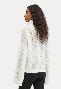 Pullover chunky in maglia a treccia di colore bianco, con colletto alto, maniche lunghe e un design a maglia aperta con una finitura testurizzata.