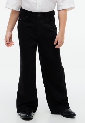 Enfant portant une chemise blanche à manches courtes, un pantalon noir à jambes larges et des chaussures noires, debout sur un fond blanc.