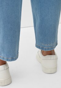 Pantalones vaqueros de corte amplio en un tenue color azul claro con una textura sutil, presentando un dobladillo con el logotipo bordado "TRIANGLE" y combinados con zapatillas blancas.