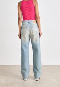 Personne portant un débardeur rose et un jean bleu clair délavé et ample, debout dos à un mur et un sol unis.