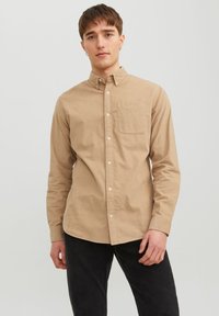 Camisa button-up de algodón marrón claro con un bolsillo. Mangas largas y cuello clásico. Llevada con pantalones oscuros, el modelo está de pie frente a un fondo sencillo.