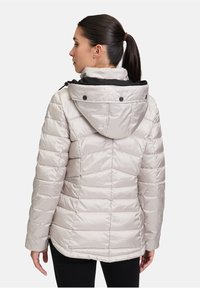 Puff jakke i lys beige med vandret og chevron quiltning. Har en aftagelig hætte, sort foring og tryklåse ved halsen.