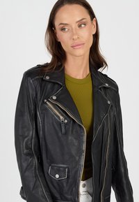 Schwarze Leder-Motorradjacke mit asymmetrischem Reißverschluss, Druckknöpfen und strukturiertem Finish. Getragen über einem grünen, figurbetonten Oberteil.