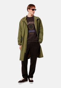 Impermeable ligero color verde oliva con capucha, superpuesto a un sudadera oscura con texto multicolor, combinado con pantalones oscuros y zapatillas marrones.