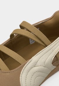 Κοντινό πλάνο καφέ παπουτσιού με τρεις ελαστικές λωρίδες και λευκό υφασμάτινο διακοσμητικό στο πλάι.