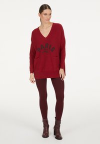 Roter Strickpullover mit V-Ausschnitt, versehen mit der Aufschrift "PARIS mon amour" in schwarzer Schrift und zwei Fronttaschen. Kombiniert mit dunkelburgunderfarbenen Leggings und Stiefeln.