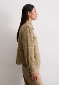 Femme aux longs cheveux noirs portant un blouson en plaid beige et une jupe assortie, se tenant de profil contre un mur clair et uni.