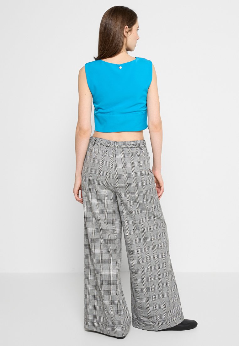 Korte, mouwloze top in helder blauw, gecombineerd met grijze plaid broek met wijde pijpen en hoge taille. Achterkant met knoopdetail zichtbaar.