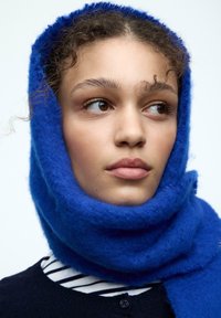 Sciarpa di lana blu che avvolge la testa, caratterizzata da una finitura morbida e lavorata a trama. Maglione a righe sotto, con un colore scuro che contrasta con la sciarpa brillante.