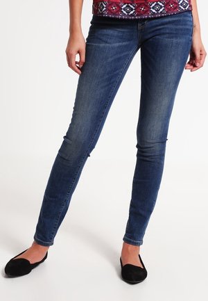 Mujer con vaqueros ajustados azules, bailarinas negras y una blusa roja con estampado, de pie con las piernas cruzadas en los tobillos.