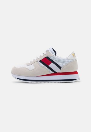 Witte en beige sneaker met rood-wit-blauwe strepen, witte veters en een rood-wit-blauwe gelaagde zool, zijaanzicht op witte achtergrond.