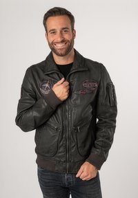 Schwarze Leder-Bomberjacke mit Reißverschluss vorne, zwei unteren Taschen und dekorativen Aufnähern. Rippbündchen an den Ärmeln und am Saum.