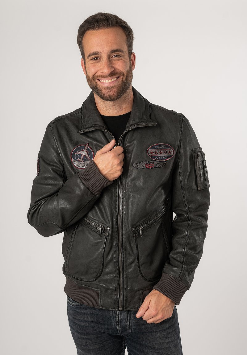 Schwarze Leder-Bomberjacke mit Reißverschluss vorne, zwei unteren Taschen und dekorativen Aufnähern. Rippbündchen an den Ärmeln und am Saum.