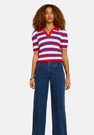 Frau mit lockigem Haar, die ein kurzärmeliges Stricktop mit rot-weiß-blauen Streifen und hoch taillierte, weit geschnittene blaue Jeans trägt, stehend vor weißem Hintergrund.