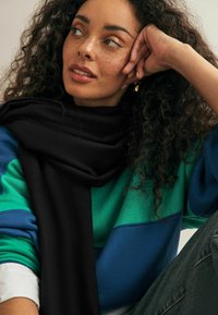 Écharpe noire en tissu doux, enroulée autour du cou. Le mannequin porte un sweat-shirt coloré en blocs de vert et de bleu, avec un jean à coupe décontractée.