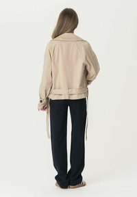 Giacca leggera beige con maniche lunghe, vita con cintura e ampio colletto, abbinata a pantaloni neri a gamba larga e sandali.