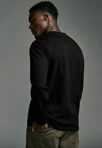 Schwarzes Langarmshirt mit glatter Textur, lockerer Passform und rundem Ausschnitt, von hinten getragen mit olivgrünen Hosen.