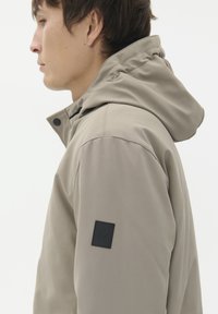 Vyras, dėvintis šviesiai smėlinį pūkinį švarką su juodu kvadratiniu logotipu ant viršutinio rankovės, pasisukęs į kairę prieš baltą fono.