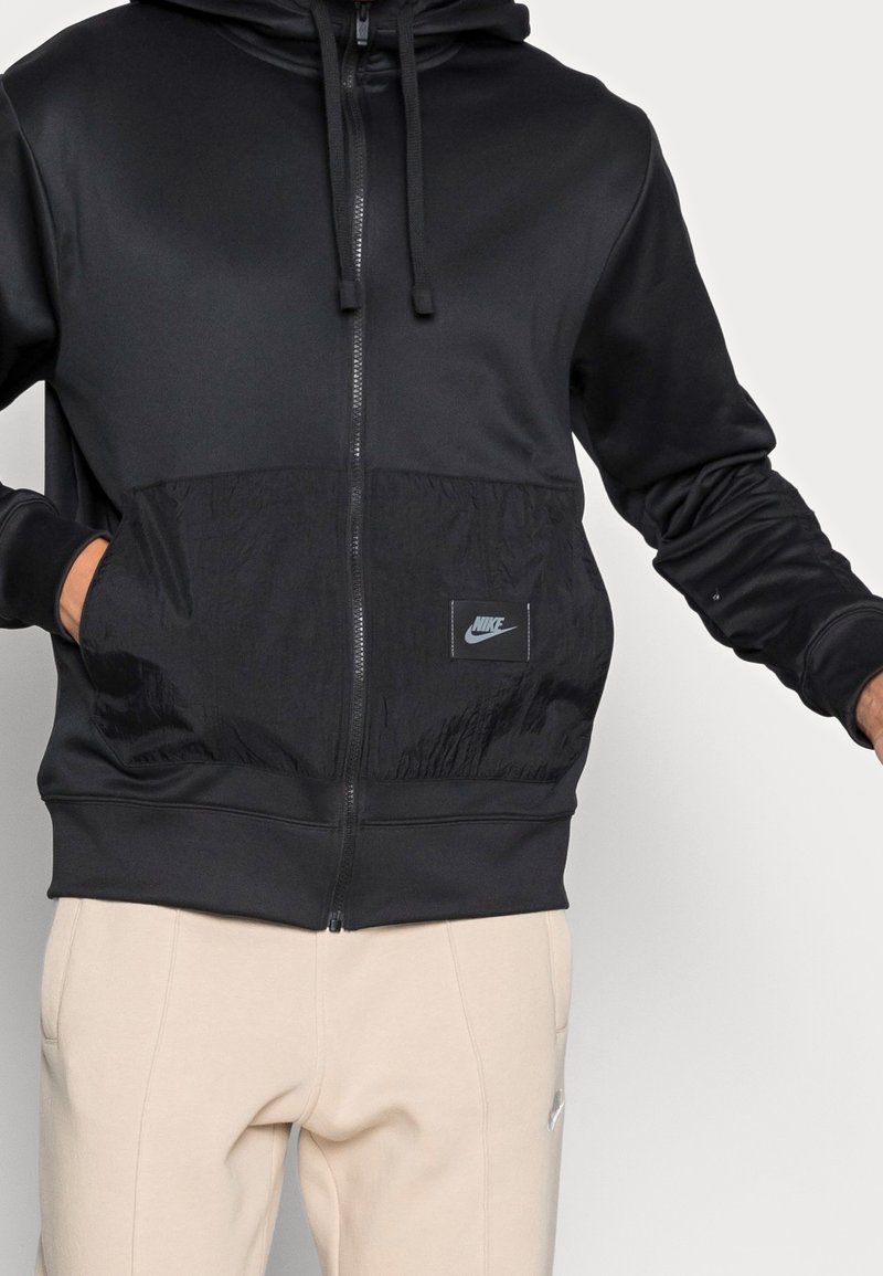 Felpa nera con zip, dotata di un pannello inferiore testurizzato e un piccolo patch con il logo Nike. Presenta un cappuccio e un cordoncino, realizzata in tessuto liscio.