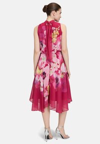 Blumenkleid in knielanger Länge in Rosa-Tönen mit fließendem Schnitt, Rückenschnürdetail und transparenter Überlagerung. Kombiniert mit silbernen High Heels.