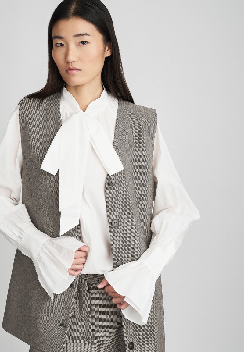 Grijze tweed vest met knopen aan de voorkant en een gecentreerde kraag, gecombineerd met een witte blouse met een strik en flared mouws.