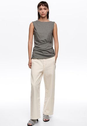 Mujer de pie con una blusa gris sin mangas con fruncidos, pantalones sueltos color blanco hueso y sandalias grises abiertas de tacón sobre un fondo blanco liso.