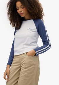 Biała i granatowa koszulka z długim rękawem, okrągłym dekoltem oraz napisem "SUPERHEAT", z granatowymi rękawami ozdobionymi trzema białymi paskami.