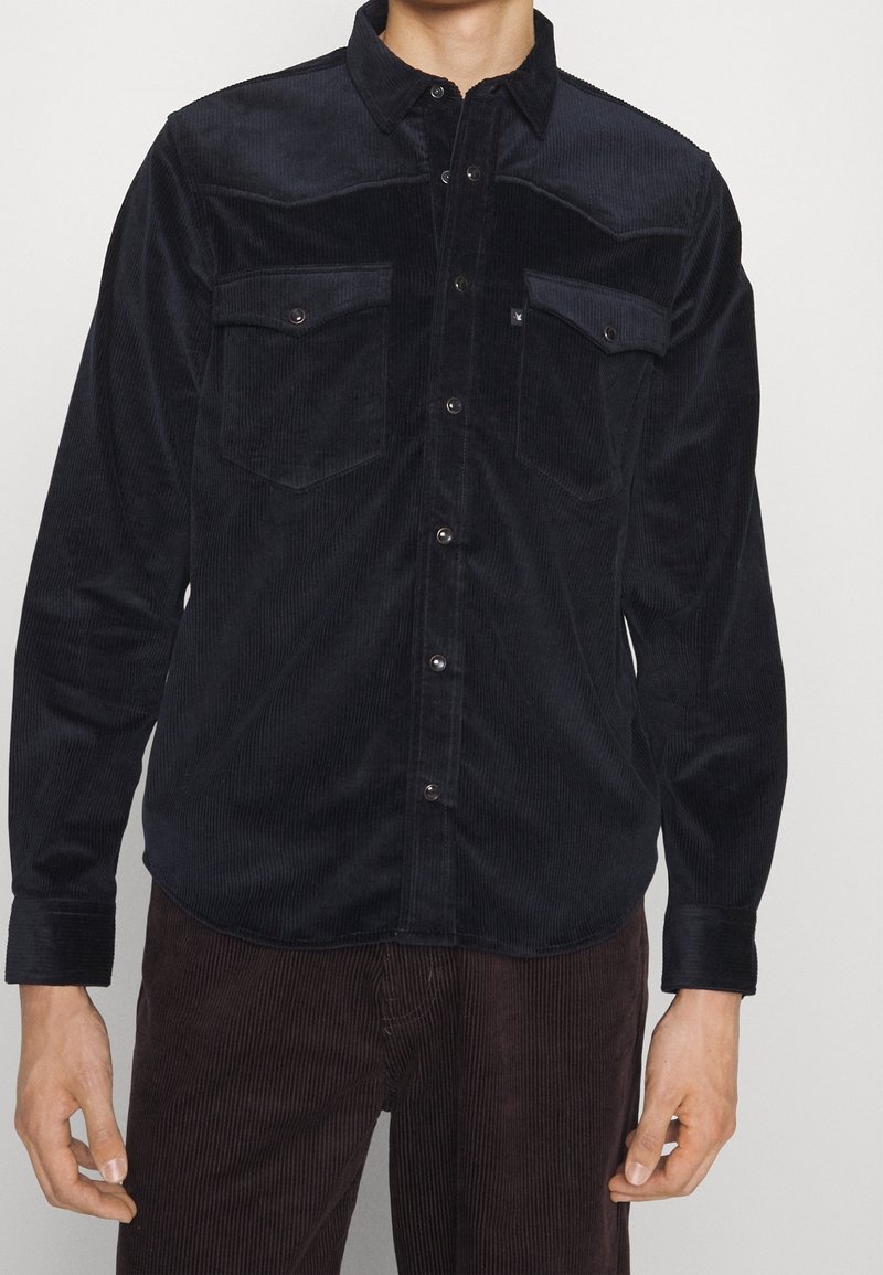 Ciemna sztruksowa koszula z zapięciem na guziki, dwoma kieszeniami na piersiach i klasycznym kołnierzykiem. Żebrowana faktura z metalowymi guzikami.