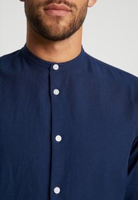 Mörkblå skjorta med mandarin-krage, som har fem vita knappar fram och ett slätt, texturerat tyg.