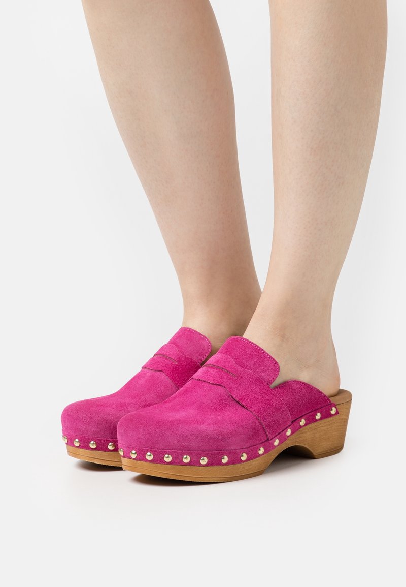 Sabots en daim rose vif à enfiler avec talons en bois et clous dorés autour de la semelle, portés pieds nus sur fond blanc.