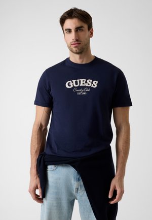 Mężczyzna w granatowym t-shircie z napisem „GUESS Country Club EST. 1981” i jasnoniebieskich dżinsach, z ciemną bluzą zawiązaną wokół talii.