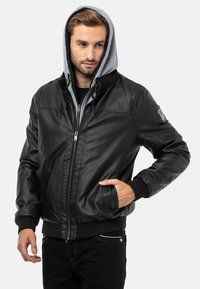 Schwarze Lederbomberjacke mit grauem Kapuze, Vorderreißverschluss, gerippten Bündchen und einem Patch am linken Ärmel. Das Model posiert mit den Händen in den Taschen.