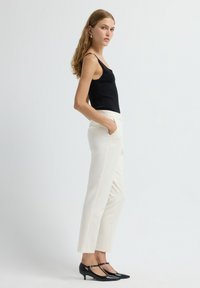 Black sleeveless top, white tailored trousers with side pockets, and black heeled shoes. Smooth fabric and fitted design, side profile view.