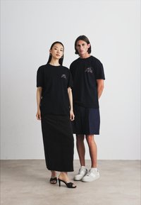 Două modele stau în fața unui fundal simplu. Femeia poartă un tricou negru și o fustă neagră, până la podea. Bărbatul poartă un tricou negru și pantaloni scurți închisi la culoare, cu adidași albi.